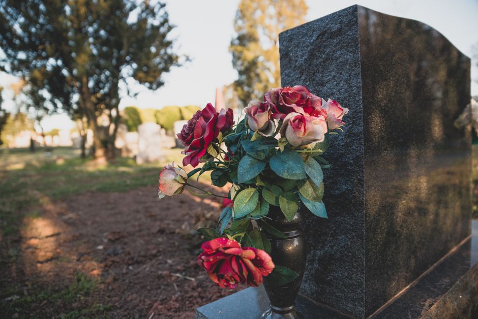 A floral tribute featuring a bouquet of artificial roses in shades of pink and red, with green foliage, arranged in a small black vase. The bouquet rests against a polished black granite gravestone in