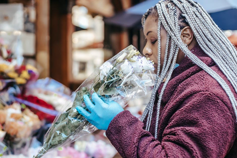 A woman with braided, silver-streaked hair wearing a maroon fleece jacket and blue gloves is holding a bouquet of white roses wrapped in transparent cellophane at an outdoor flower market. She is clos