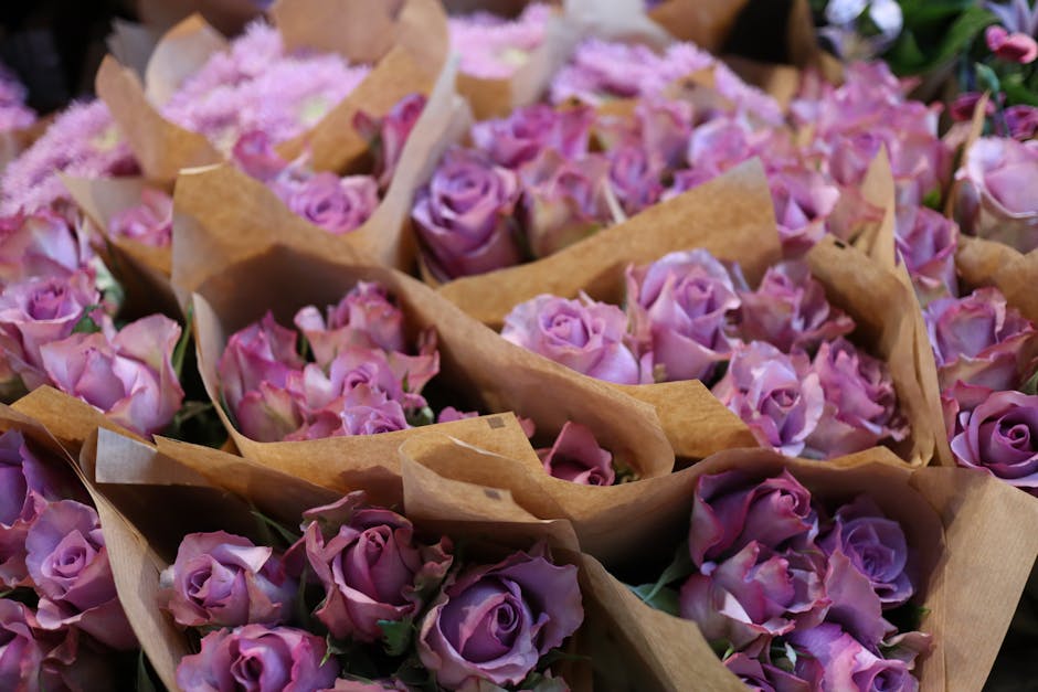 A collection of fresh pink and lavender roses arranged in small brown kraft paper wraps, ready for flower delivery. The roses have partially opened petals with a velvety texture, accompanied by green